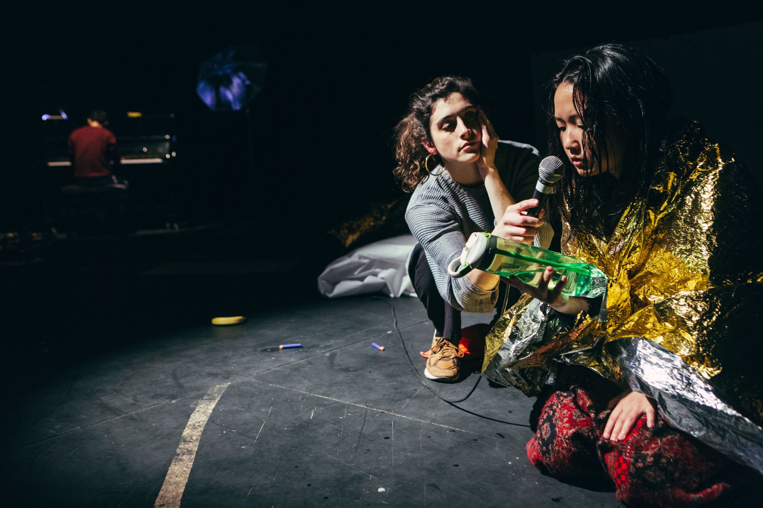 photo of a girl on stage wearing a thermal blanket and holding a plastic bottle, while another girl holds a microphone to her mouth