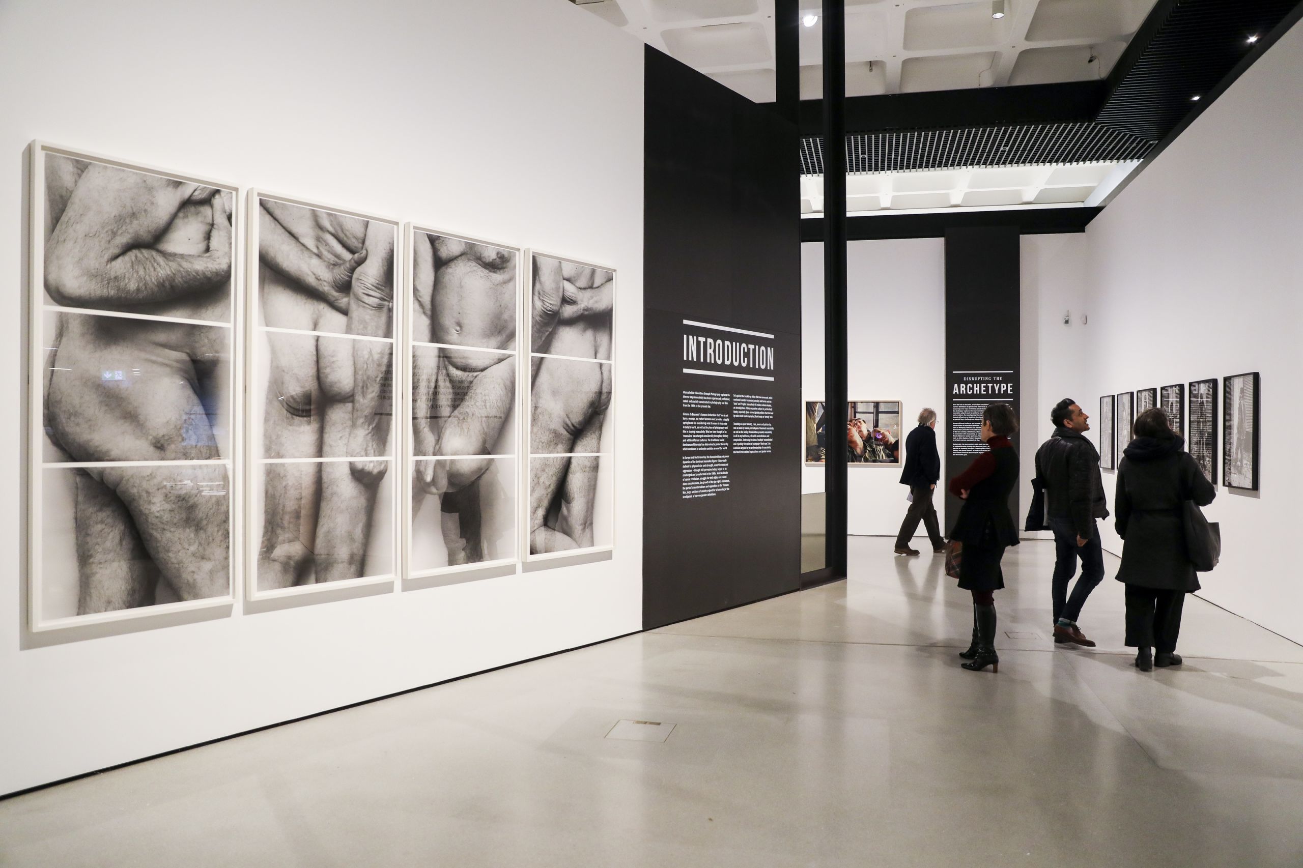 People stand in a white gallery space looking photographs hung on the wall, a large black and white photographic image is displayed, showing the nude figure of an elderly man.