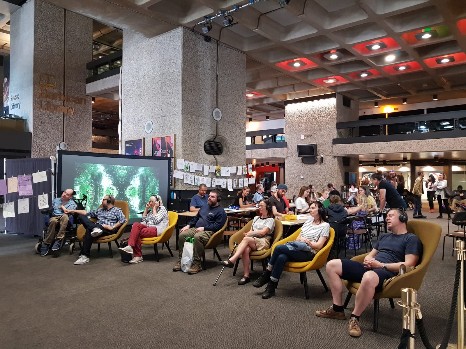 A group of people sit on yellow chairs wearing headphones, watching a large television screen