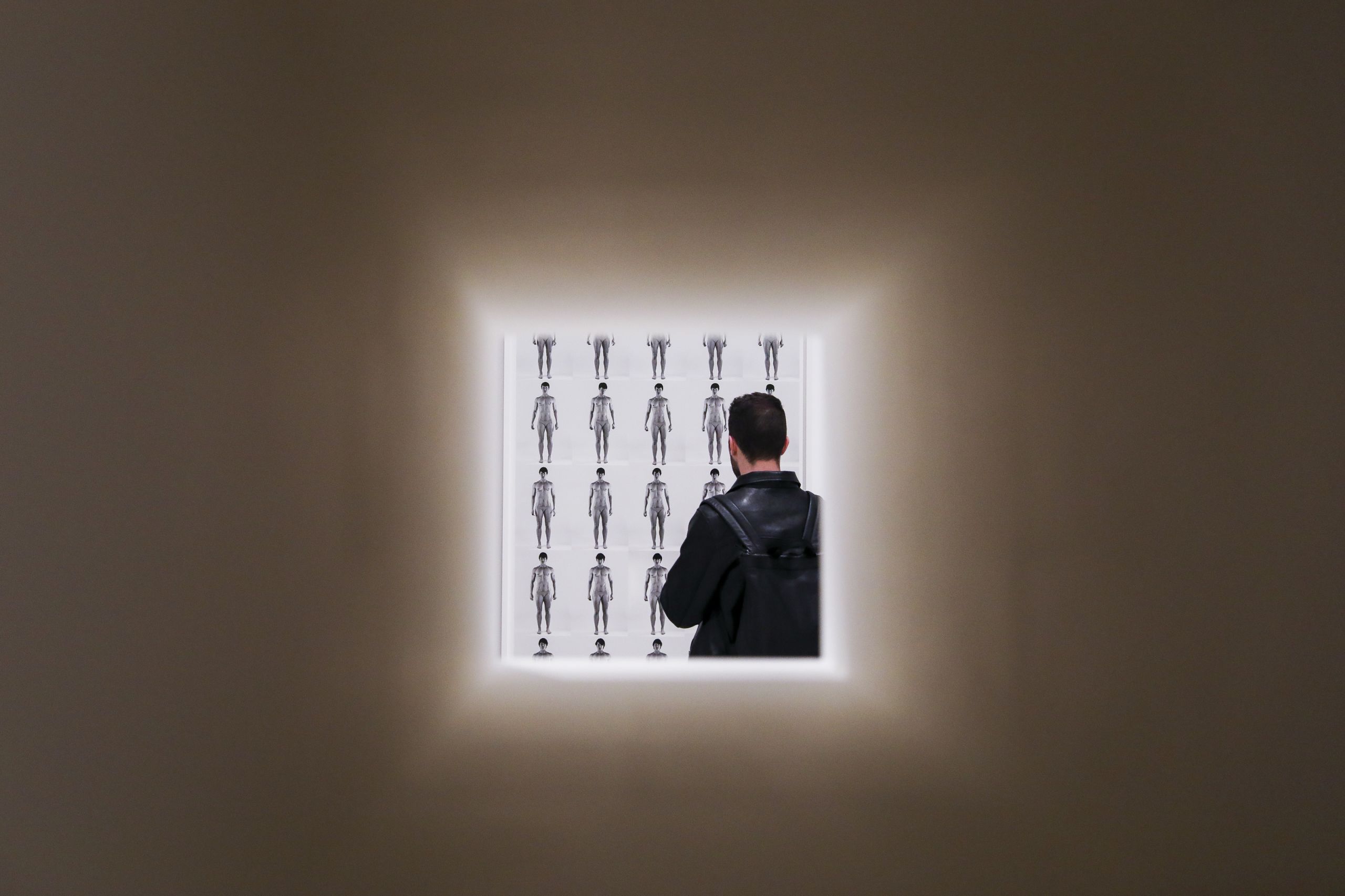 A man in a black jacket with a backpack looks at a black and white image featuring a grid of timelapse photographs of a muscular figure. The image is framed in a square, with the photograph taken through a hole in the gallery wall. 