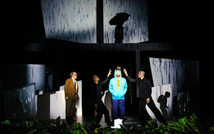 A man in a blue hoodie stands in the middle of the stage surrounded by film projections of rain